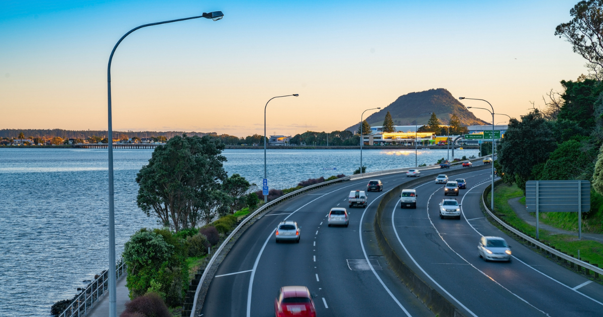 Image of cars driving on the road with Mount Maunganui in the background, showing the kinds of views you can see if you are moving to NZ and need to buy a car