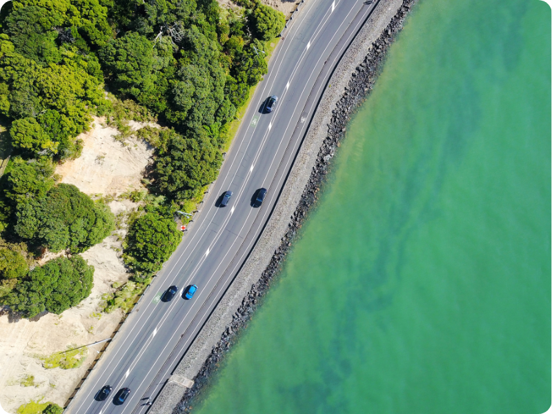Moving to NZ and looking to buy a car as a foreigner, we can make it easy. Image of cars driving along a road in NZ with ocean one one side